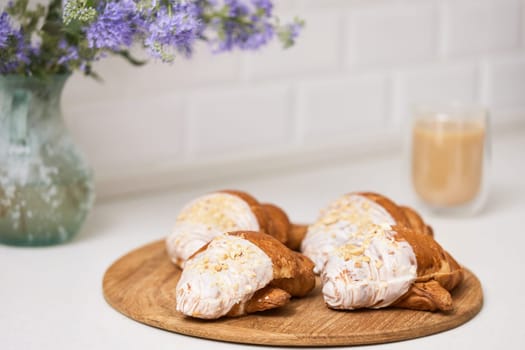 Delicious fresh croissants on wooden tray and glass of coffee on white table, closeup, tasty breakfast