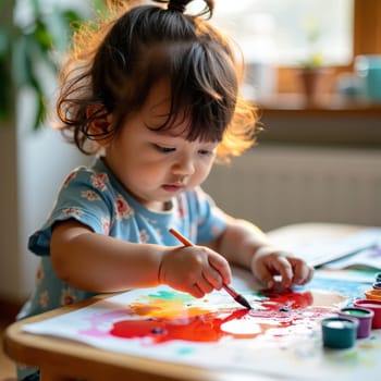 Young Asian girl in floral shirt concentrating on colorful finger painting, showcasing childhood creativity and artistic expression in a sunlit home setting.