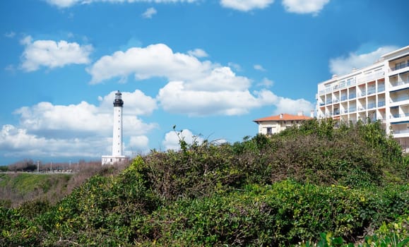 A Biarritz lighthouse is on a hill next to a building. The sky is blue with some clouds. The scene is peaceful and serene