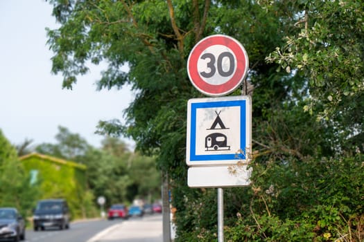 A french sign with a red circle and number 30 . The sign is surrounded by trees and is near a road with cars