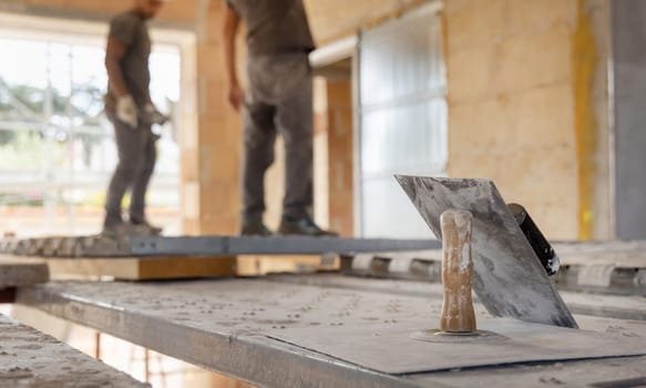 Craftsmen working inside a room standing on scaffoldings, metal trowels in the foreground. Men in grey work clothes during construction or renovation process