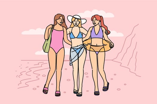 Women in bikinis walk along beach on seashore, enjoying summer holiday at hot resort. Three tourist girls pose on sandy beach, inviting you to visit tropical island to sunbathe and swim
