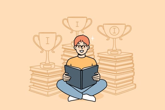 Boy prodigy elementary school student holding laptop, sitting near stacks of books with winning cups. Happy schoolchild prodigy who won online olympiad thanks to reading educational literature