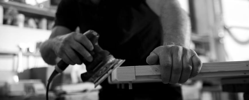 Black and white image of A carpenter is sanding a piece of wood using an electric sander in his workshop