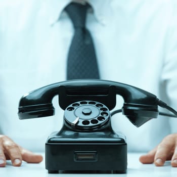 Office worker in a white shirt and tie is sitting at a desk with his hands resting in front of a vintage black telephone
