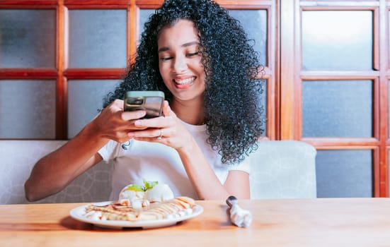 Beautiful girl sitting photographing a chocolate crepe and ice cream. Smiling woman taking a photo of a chocolate crepe on table