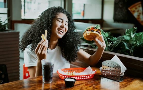 Happy girl enjoying a hamburger in a restaurant. Afro young woman enjoying a hamburger with fries in a restaurant.