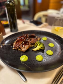 Sophisticated dish features tender slices of duck garnished with broccoli and artistically arranged green sauce dollops on a black ceramic plate. Elegance and precision of modern culinary presentation in a fine dining setting.