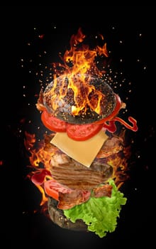 Cheeseburger with a piece of meat, tomato, green lettuce and a bun with sesame seeds on a black background with fire