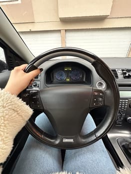 Female hand on a steering wheel inside a modern car, showcasing a casual outfit with a fluffy jacket and jeans. Moment just before starting drive, highlighting dashboard and residential garages outside.