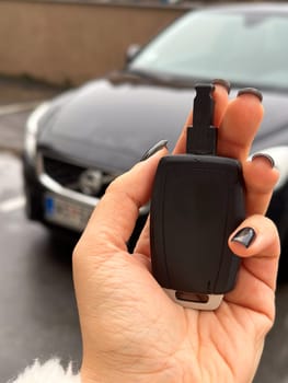 Woman's hand holding a car key fob in front of a black modern car on a rainy day. Fluffy jacket highlights the close-up shot of the key, ready to unlock the vehicle