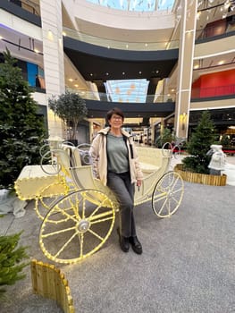 Woman in casual attire standing next to a decorative carriage in a modern shopping mall. Christmas holiday decorations and festive atmosphere create a lively and cheerful scene.