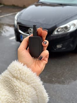 Woman's hand holding a car key fob in front of a black modern car on a rainy day. Fluffy jacket highlights the close-up shot of the key, ready to unlock the vehicle