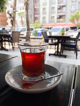 Glass of tea on a white saucer with a spoon, set on an outdoor cafe table with a view of a bustling urban street scene in the background. Essence of a relaxing tea moment amidst city life.