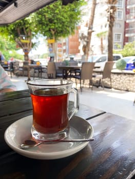 Glass of tea on a white saucer with a spoon, set on an outdoor cafe table with a view of a bustling urban street scene in the background. Essence of a relaxing tea moment amidst city life.