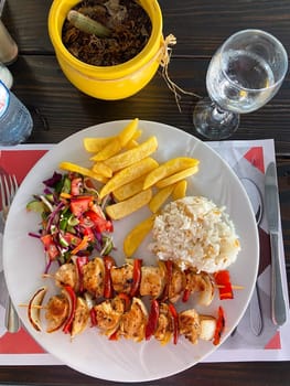 Delicious plate of grilled chicken skewers with rice, salad, and fries on a wooden table setting. Well-balanced and colorful meal, perfect for a vibrant dining experience.