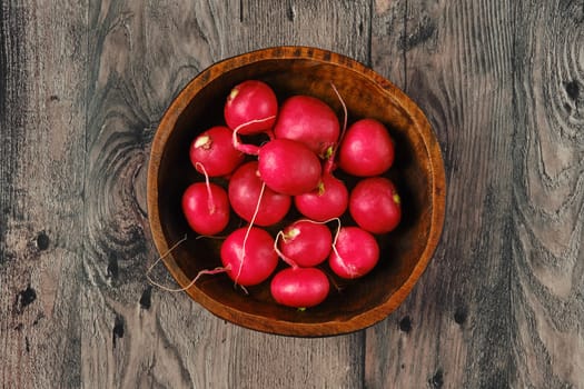 A rustic wooden bowl with vibrant red radishes on a textured, weathered wooden table. The fresh produce showcases the natural beauty of farm-fresh vegetables ready for cooking.