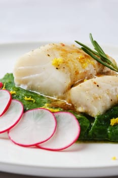 A tender white fish fillet is elegantly placed atop vibrant wilted greens. Fresh radish slices and citrus zest add a burst of color and flavor, creating a refined culinary presentation.