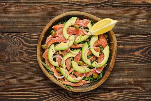 Salad with slices of fresh salmon, creamy avocado, and crisp onions, artfully arranged in a wooden bowl. It is complemented by a wedge of lemon, adding a refreshing touch.