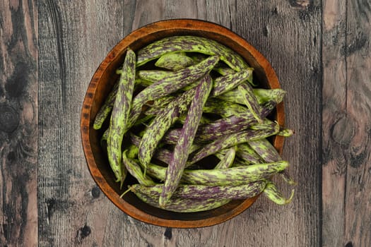 A wooden bowl filled to the brim with vibrant purple and green beans rests on a textured wooden surface. Dragon tongue beans in a bowl.