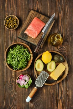 An assortment of fresh ingredients on a wooden surface, including salmon fillet, avocado, lemon, and various herbs, ready for assembling a vibrant salad.