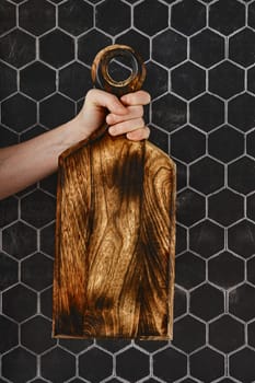 A person grips a stunning wooden cutting board with intricate grain patterns, showcasing craftsmanship. The board is held up against a modern black hexagonal tile wall, adding a stylish touch.