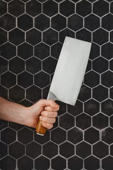 A hand grips a large kitchen cleaver, positioned against a striking hexagonal tile backdrop. The wooden handle adds a touch of warmth to the contemporary kitchen environment.