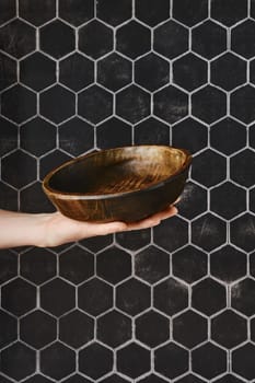 A hand presents a beautifully crafted wooden bowl, showcasing its smooth finish and unique design. The bowl contrasts with a striking hexagonal tile wall, adding elegance to the kitchen setting.