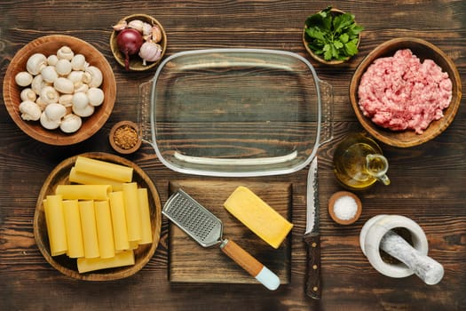 Fresh ingredients are arranged neatly on a wooden countertop in preparation for a pasta meal. Components include mushrooms, ground meat, pasta sheets, cheese, garlic, and herbs.