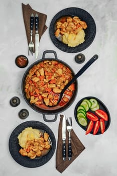 A hearty chicken or pork stew in a black plates, accompanied by creamy mashed potatoes, fresh tomato, and cucumber slices on a marble dining tabletop, perfect for a comforting meal shared with family