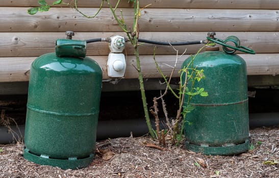 Two green gas tanks are sitting on the ground next to a wooden wall. The tanks are connected to a gas line