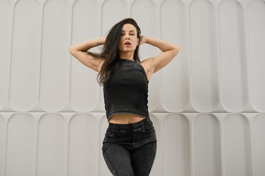 Woman holding both hands behind her head while standing against a white wall