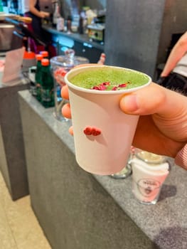 Close-up hand holds a cup of vibrant green matcha in a cozy cafe setting, Freshness of beverage. On background reveals a glimpse of the barista's workspace, filled with colorful straws and utensils