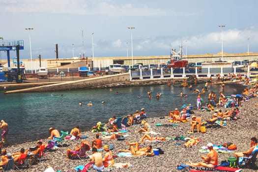Puerto de las Nieves, Gran Canaria Island, Spain - 20 July 2024. tourists on the stony beach Playa de las Nieves with a view of the harbour.