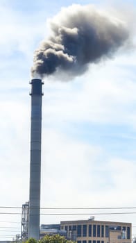 Tall industrial smokestack releasing dark smoke into the blue sky, illustrating environmental concerns and industrial emissions in an urban setting.