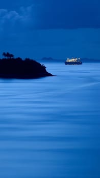 Serene nighttime seascape with an illuminated ship sailing between a silhouetted island and distant mountains, creating a tranquil tropical atmosphere.