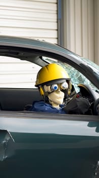 A crash test dummy wearing a yellow helmet sits in a car with a shattered windshield, simulating the aftermath of a collision for safety testing.