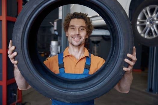 Portrait of funny smiling auto mechanic looking through car tire standing over automotive garage station interior