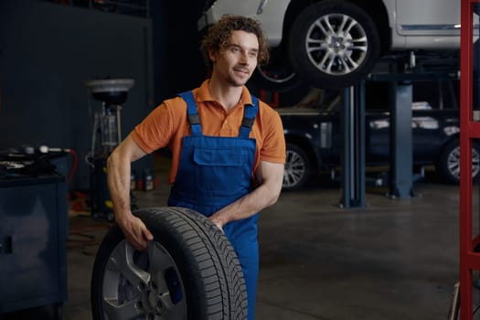 Portrait of smiling male auto mechanic carrying car tire. Professional wheel replacement and fixing service at repair shop concept