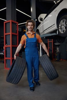 Full-length portrait of smiling male auto mechanic carrying car tires. Professional wheel replacement and fixing service at repair shop concept