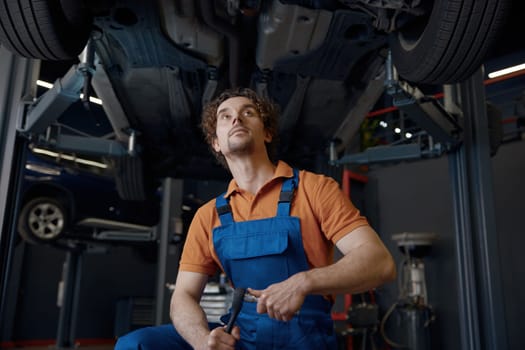 Serious and concentrated man car mechanic holding wrench tool looking at bottom of lifted automobile at repair service station
