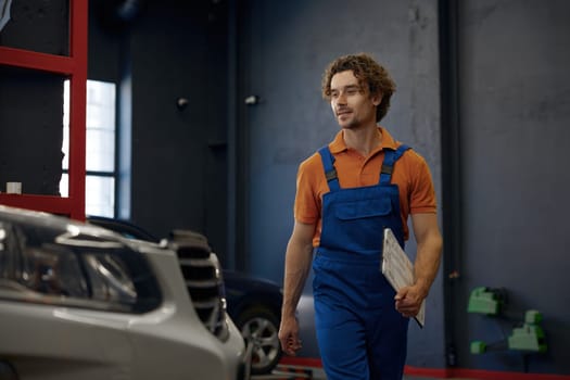 Smiling repairman car technician walking with clipboard in hands ready to start doing diagnostic, inspection and fixing problem of custom car at auto service