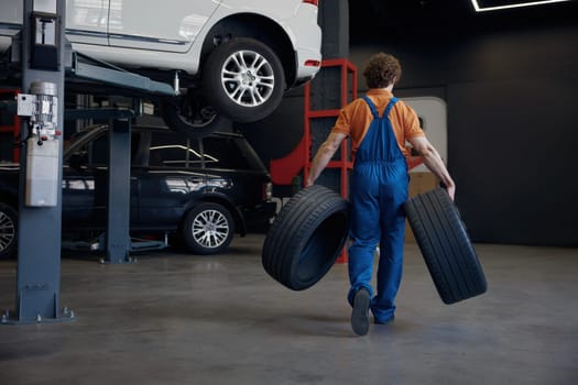Full-length shot of male auto mechanic carrying car tires. Professional wheel replacement and fixing service at repair shop concept