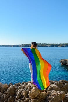 Person on rocky shore holding a colorful rainbow flag against a clear blue sky and ocean backdrop, Woman, middle aged, outdoors waving pride flag, concept of libertat, equality. mallorca, balearic islands, spain