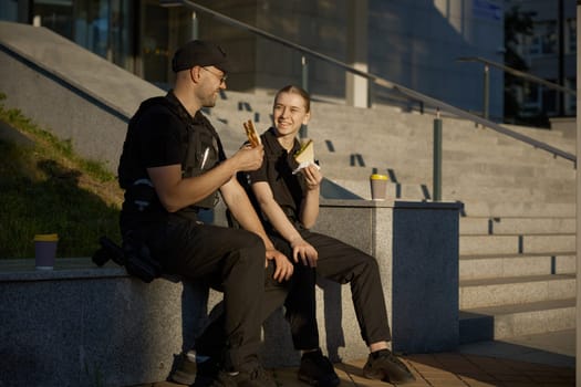 Smiling male and female patrol police officers enjoying lunch break. Policeman and policewoman resting outdoor, drinking coffee, eating sandwich and having friendly conversation