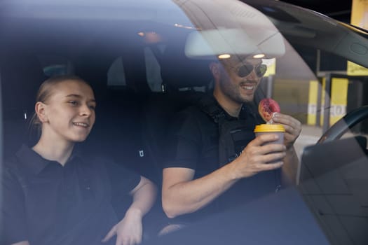 Patrol police officers diverse team sitting in car having coffee break time at work. Female colleague treating coworker with donut and hot aroma drink