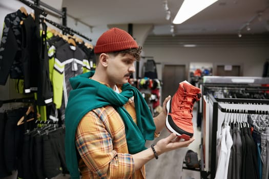 Young hipster man choosing sports sneakers shoes at sports shop store. Male customer standing with boot on hand front of camera, selective focus. Professional athlete gear at sport retail market