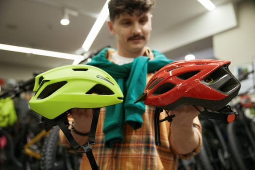 Thoughtful young hipster man choosing between two safety helmet of different color for speed bicycle riding during shopping buying gift for girlfriend at sport shop