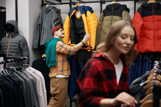 Man and woman customers choosing sportive outwear at fashion sports clothing boutique. Focus on male shopper choosing jacket on clothing hangers rack