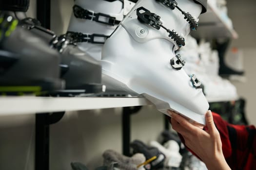 Ski boots new arrival collection on wall stand rack at professional sportive shop. Closeup view on woman hands taking white footgear accessory choosing best one